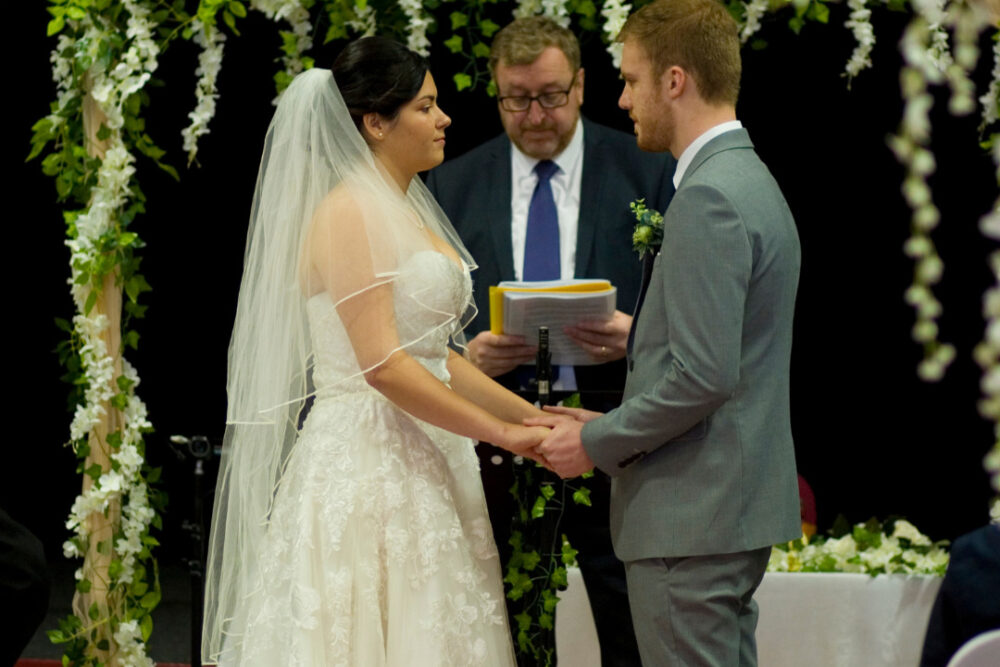 Bride in white gown and groom in grey suit holding hands, with officiant in the back